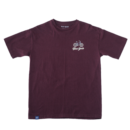 T-shirt color vino con ilustración de paisaje tropical en tonos suaves, colgada en entorno natural.